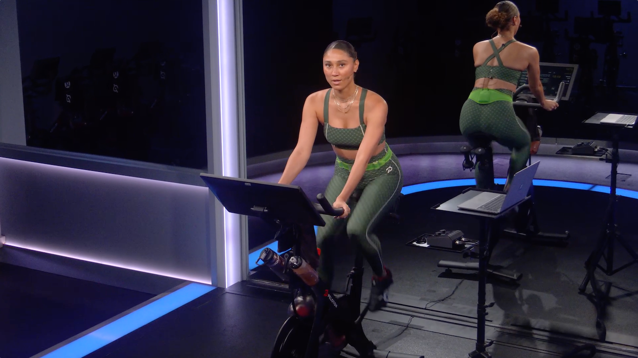 A screenshot of a Peloton instructor speaking directly into the camera while riding a stationary bike.